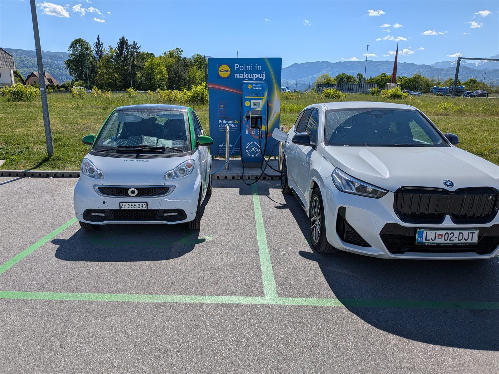 The smart ED charging at a Lidl near Bled next to a white BMW iX1, Alps in the background