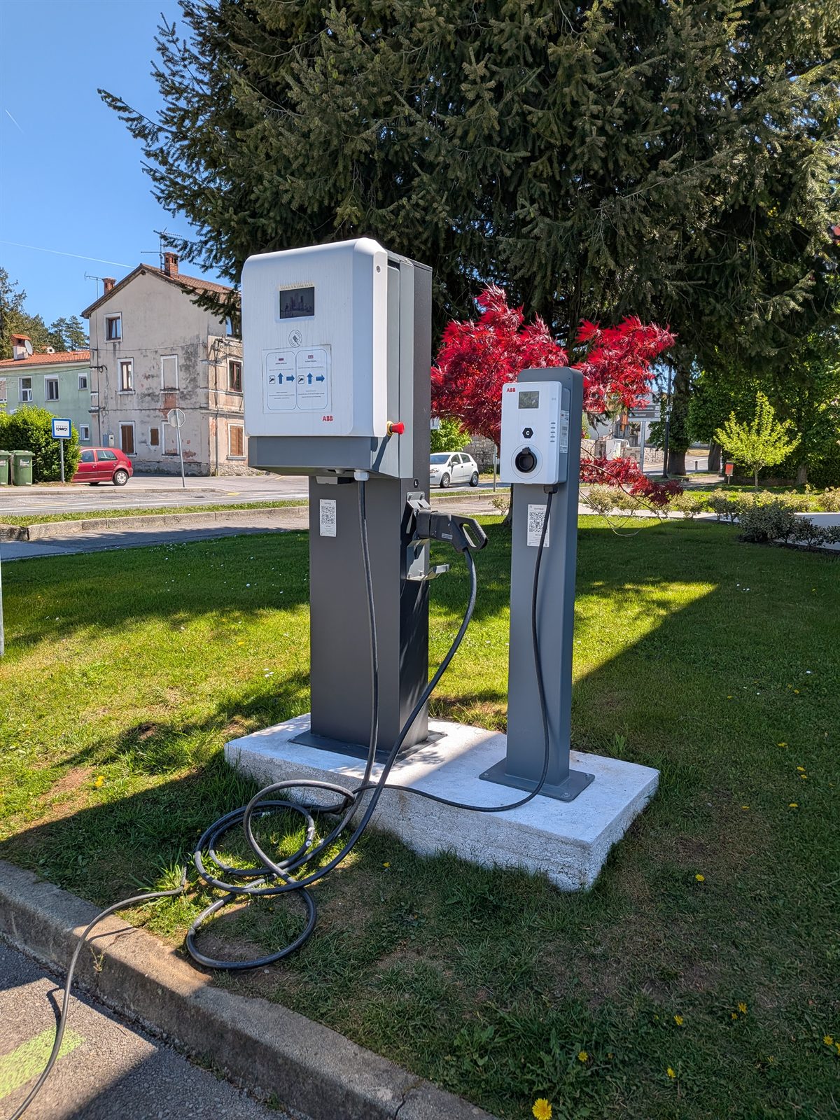 Two AC charging stations on a grass island in a Slovenian village, red maple tree behind